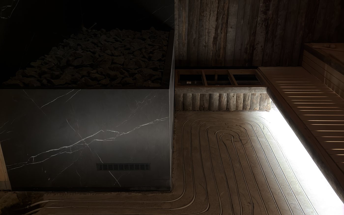 Gebogen freessleuven van de vloerkoeling in de sauna, naast de zwarte natuurstenen saunakachel